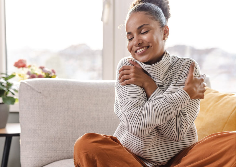 woman sitting cosy on a couch smiling while giving herself a hug